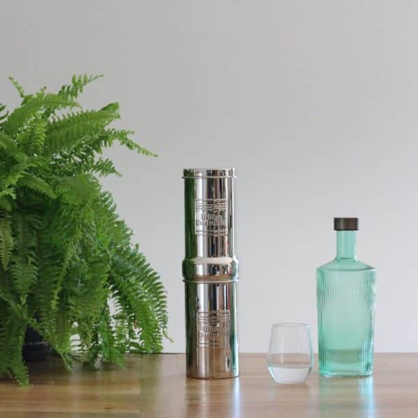 Filtre à eau British Berkefeld 1L en inox posé sur une table en bois à côté d'une plante, d'un verre et d'une bouteille Paveau Bondi